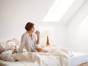 Eine junge Frau mit Nachthemd sitzt im Haus am Morgen auf dem Bett und trinkt Wasser