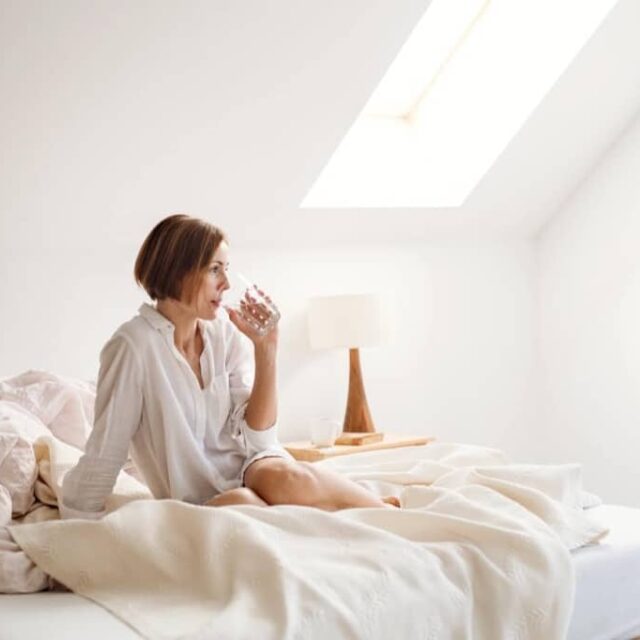 Eine junge Frau mit Nachthemd sitzt im Haus am Morgen auf dem Bett und trinkt Wasser