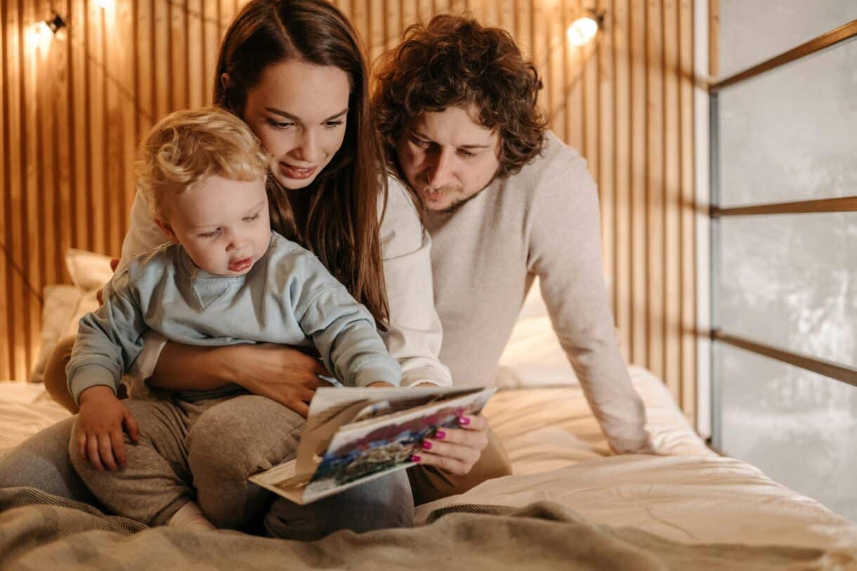 Ein Mann und eine Frau lesen ihrem Kind eine Geschichte vor - sie sitzen dabei auf einem Bett und es sieht gemütlich aus